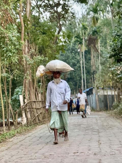 Elder walking barefoot on a village road carrying a sack